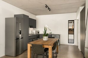 Uma cozinha moderna com geladeira de aço inoxidável, uma mesa de jantar de madeira com quatro cadeiras e uma orquídea em vaso sobre a mesa.