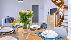 Un coin repas avec une table en bois dressée pour quatre personnes, un vase de fleurs blanches et un escalier sur le côté droit.