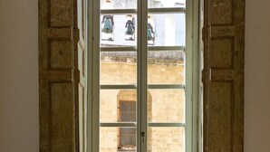 Blick durch ein Fenster mit Holzrahmen, das ein Steinhaus mit zahlreichen Fenstern und einem Glockenturm zeigt.