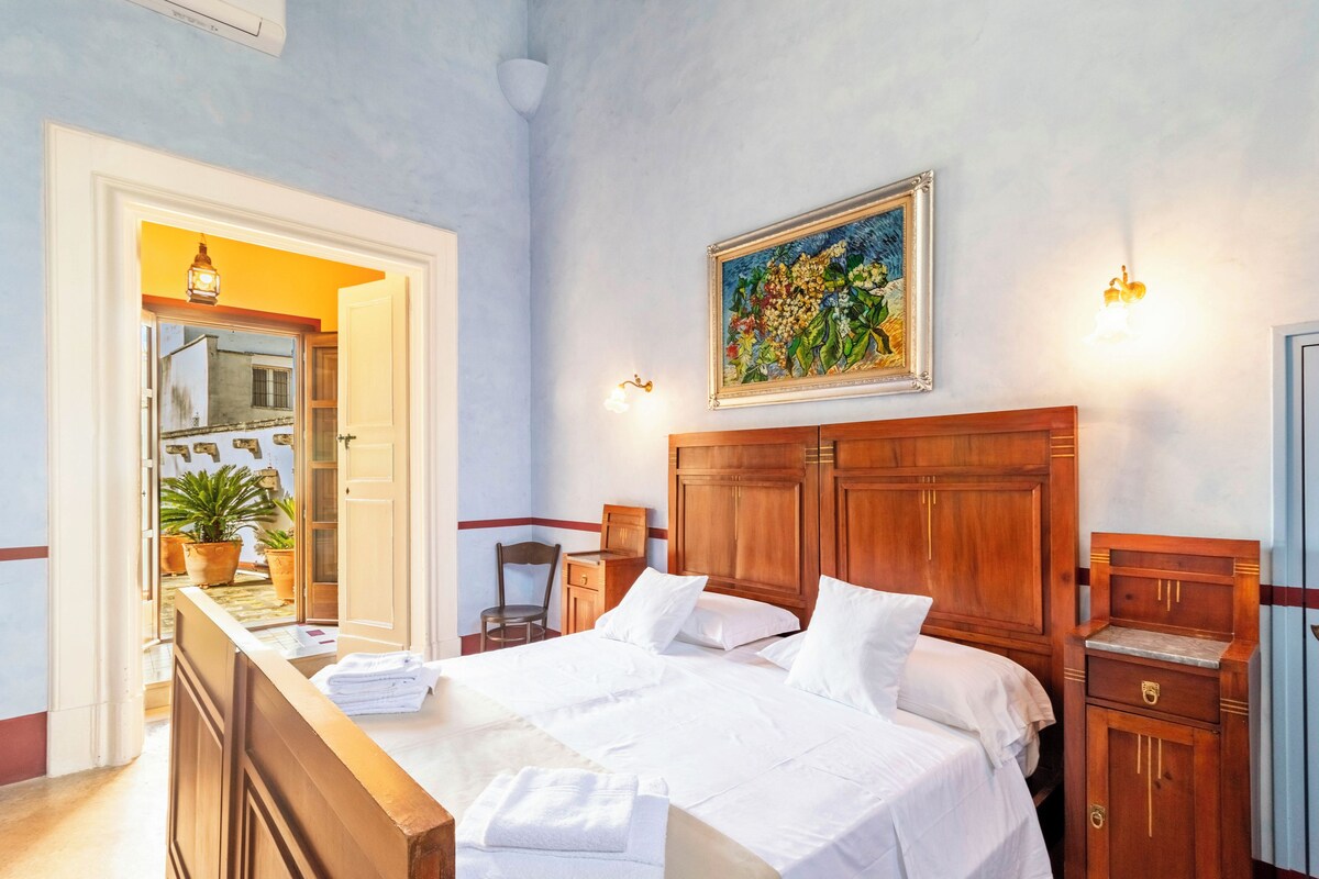 A hotel room with a large bed, wooden headboard, a painting on the wall, and a desk with a chair.