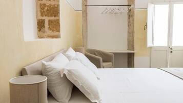 Una cama bien hecha con ropa blanca, un sofá color crema, una mesita y un perchero.