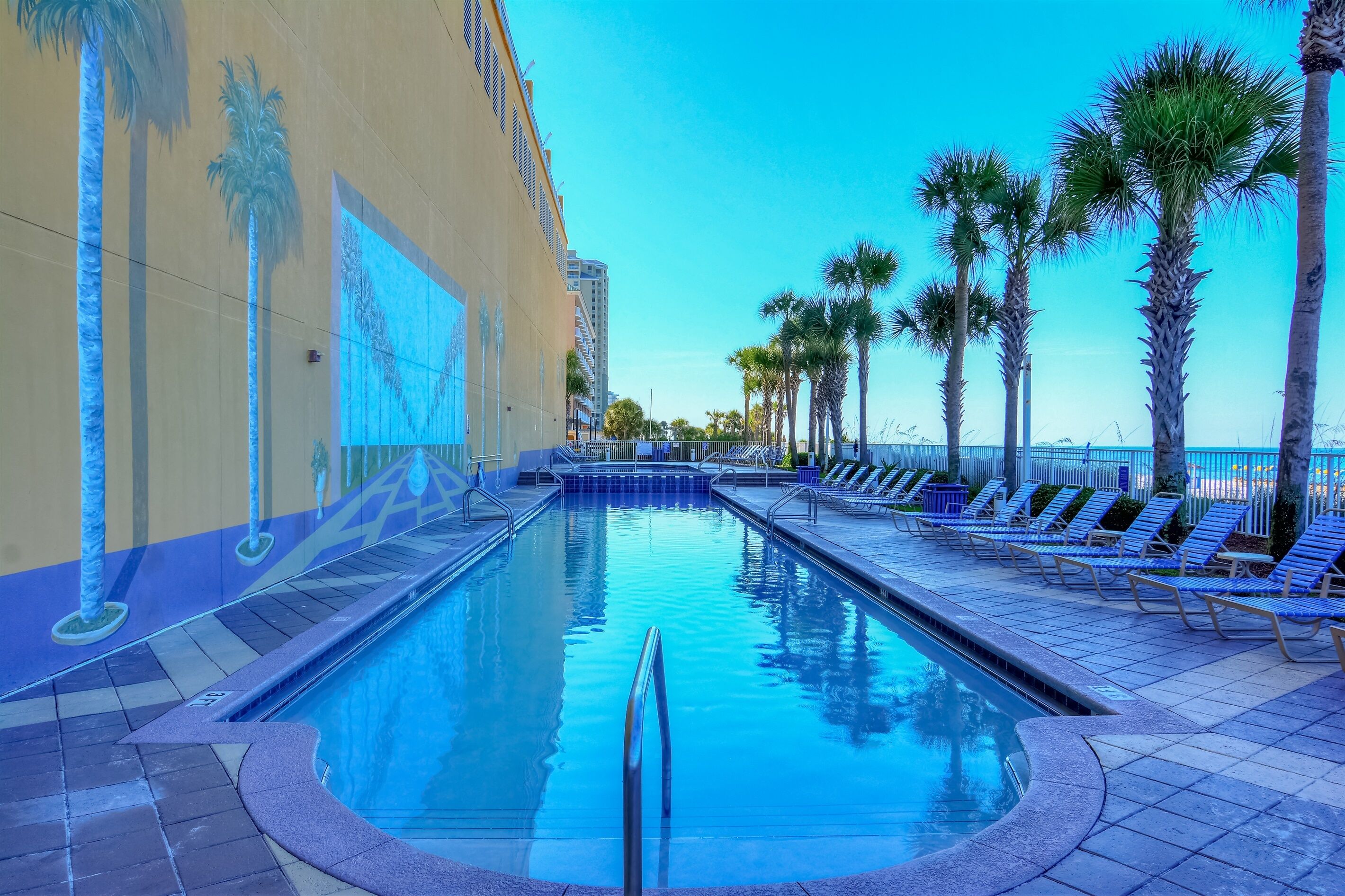 A swimming pool with a blue-tiled deck, palm trees, and lounge chairs.
