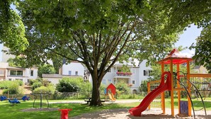 A playground with a red slide and swings, surrounded by a wooden fence and a large tree.
