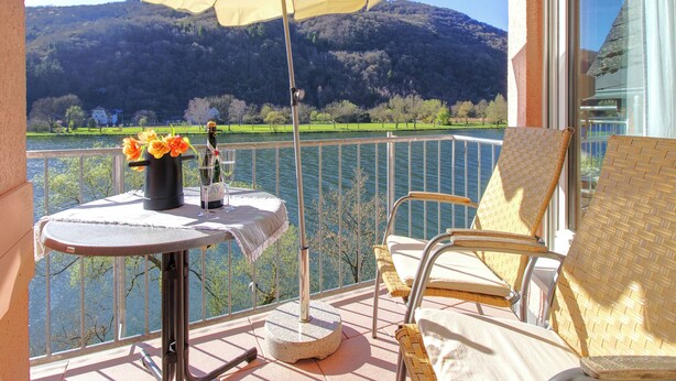 A balcony with a table, chairs, and an umbrella overlooking a lake with trees and hills.