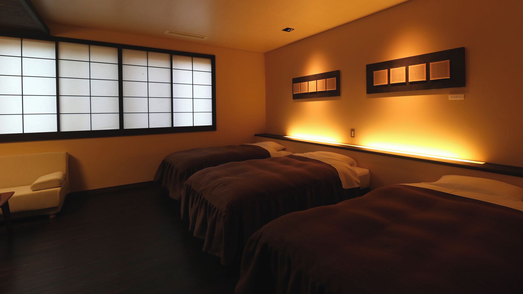 A hotel room with a traditional sliding door, two beds with dark bedding, a small sofa, and wall-mounted lighting.