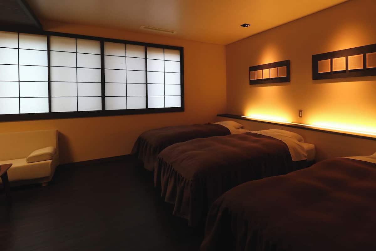 A hotel room with a traditional sliding door, two beds with dark bedding, a small sofa, and wall-mounted lighting.