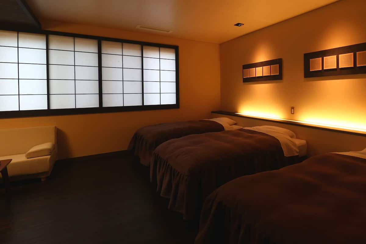 A hotel room with a traditional Japanese shoji screen, two beds with dark bedding, a small sofa, and wall-mounted lighting.