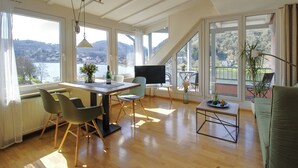 A spacious room with a dining area, a TV, and large windows offering a view of a lake and hills. - Apartment with balcony directly on the banks and with a view of the Moselle (Bullay)