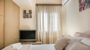 Ein Hotelzimmer mit einem Bett, einem Fernseher auf einem Nachttisch aus Holz, einem Fenster mit Vorhängen und einem an der Wand hängenden Kunstwerk.