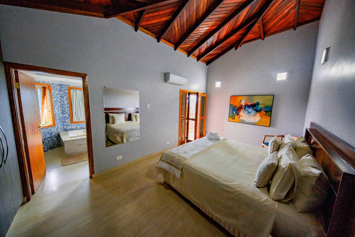 A bedroom with a wooden ceiling, a large bed, a painting, and a bathroom visible through an open door.