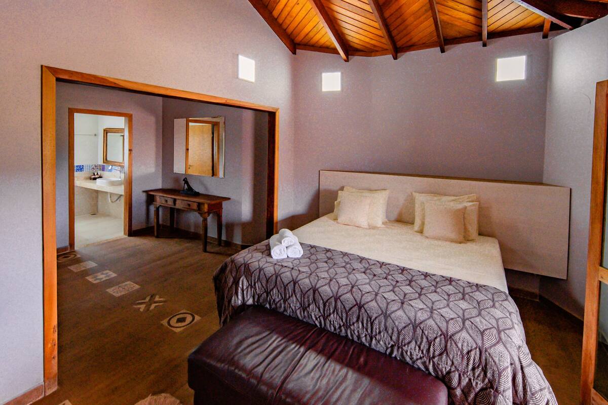 A bedroom with a large bed, a wooden ceiling, a desk, and a bathroom visible through an open door.