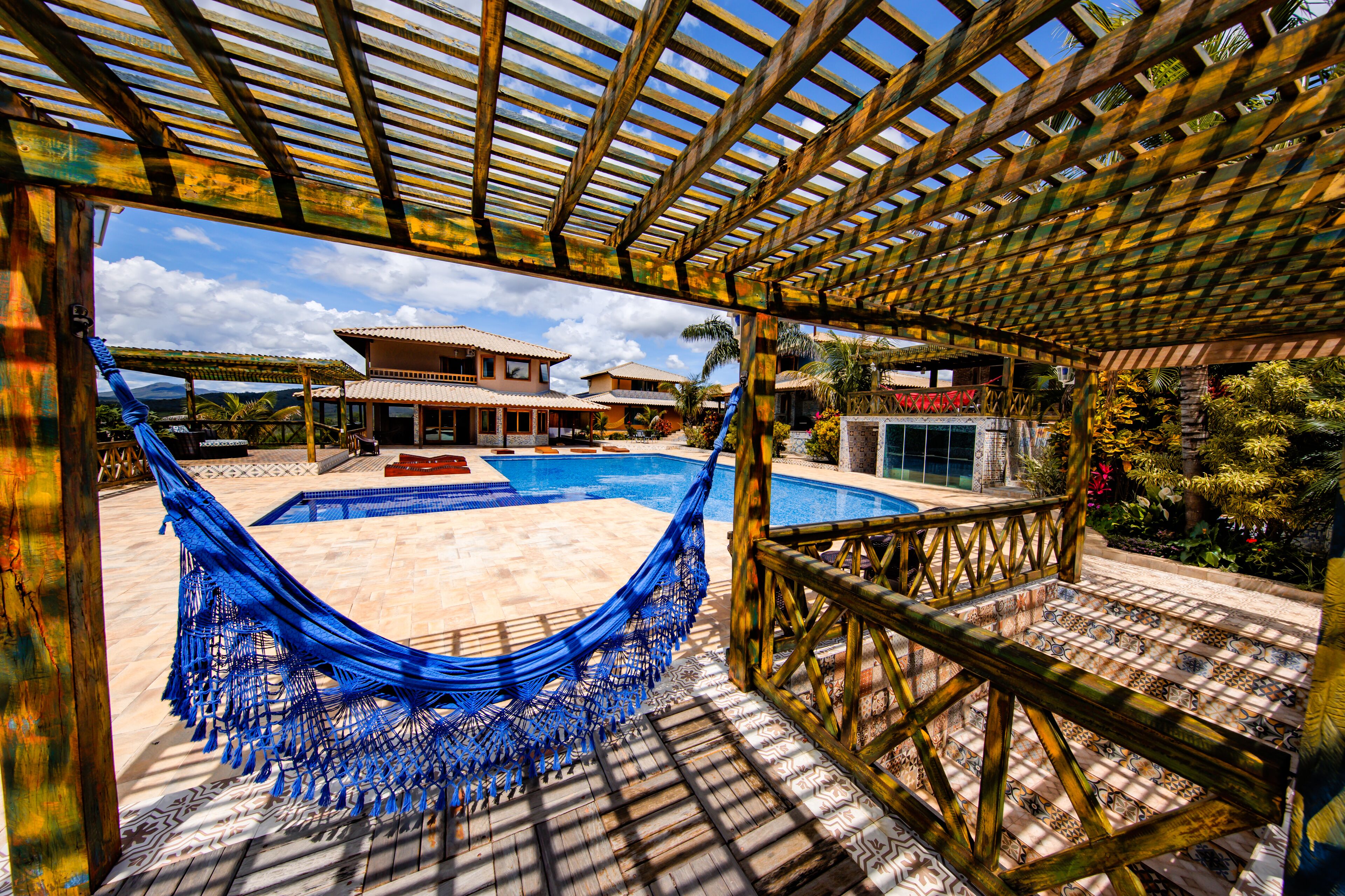 A hammock under a wooden pergola overlooking a swimming pool and a house with a tiled roof.