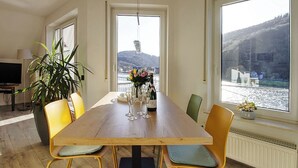 A dining area with a wooden table, yellow chairs, a potted plant, and a view of a river and mountains through large windows.