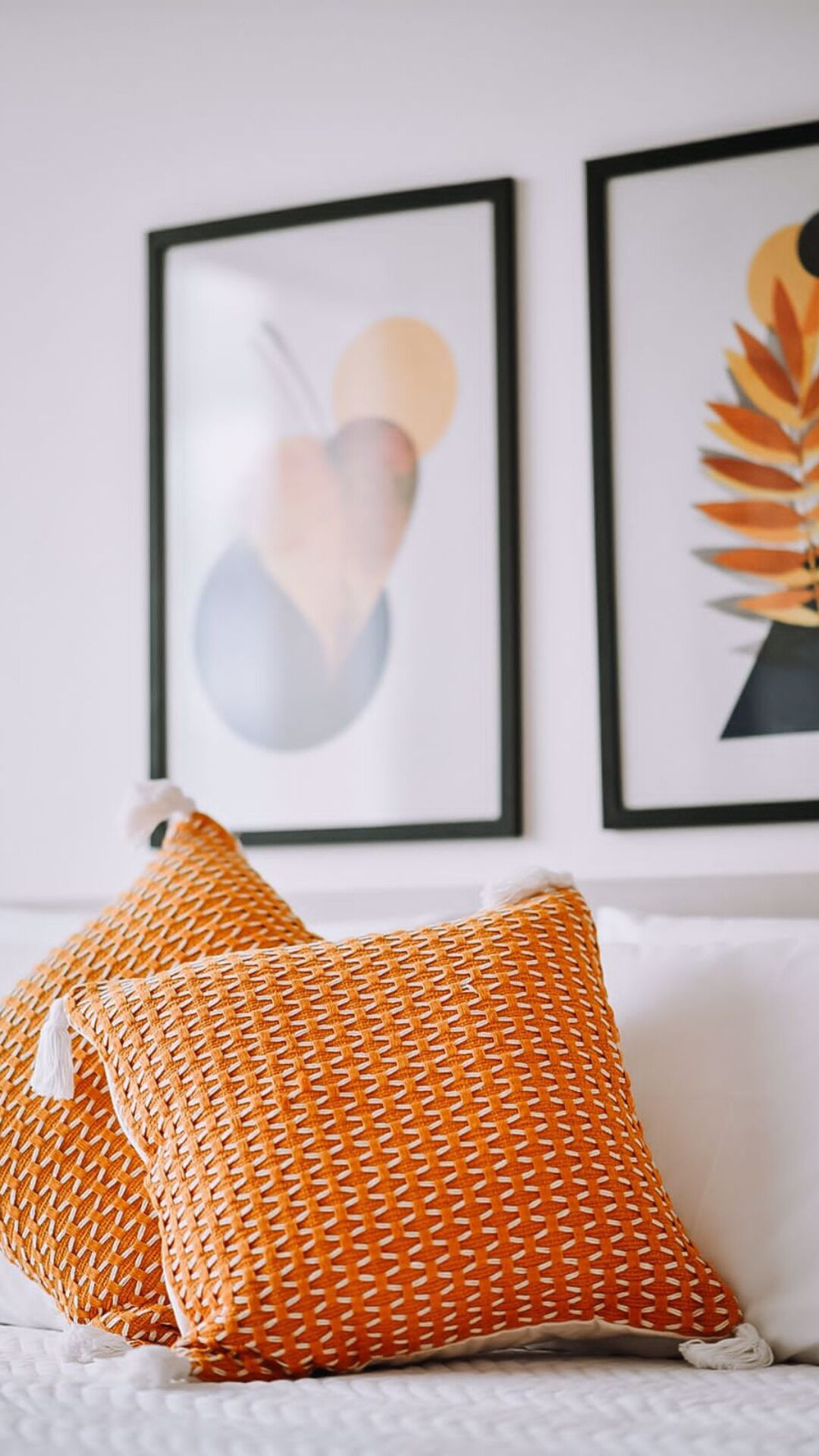 Una habitación con una cama blanca, dos almohadas naranja con estampado y dos cuadros en la pared.