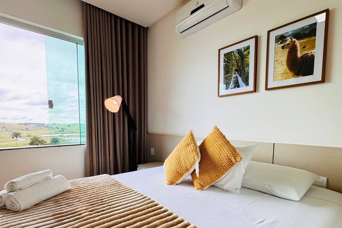 Una habitación de hotel con una cama, una ventana con cortinas, un aire acondicionado montado en la pared y dos cuadros enmarcados.
