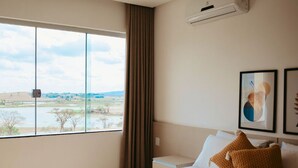 Habitación de hotel con un ventanal grande, una cama con ropa blanca, un cojín marrón y un aire acondicionado de pared.