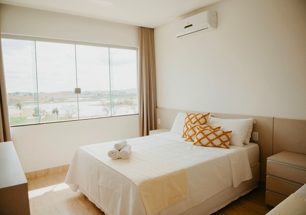 A hotel room with a large window, a bed with white bedding and an orange pillow, a bedside table, and a nightstand.