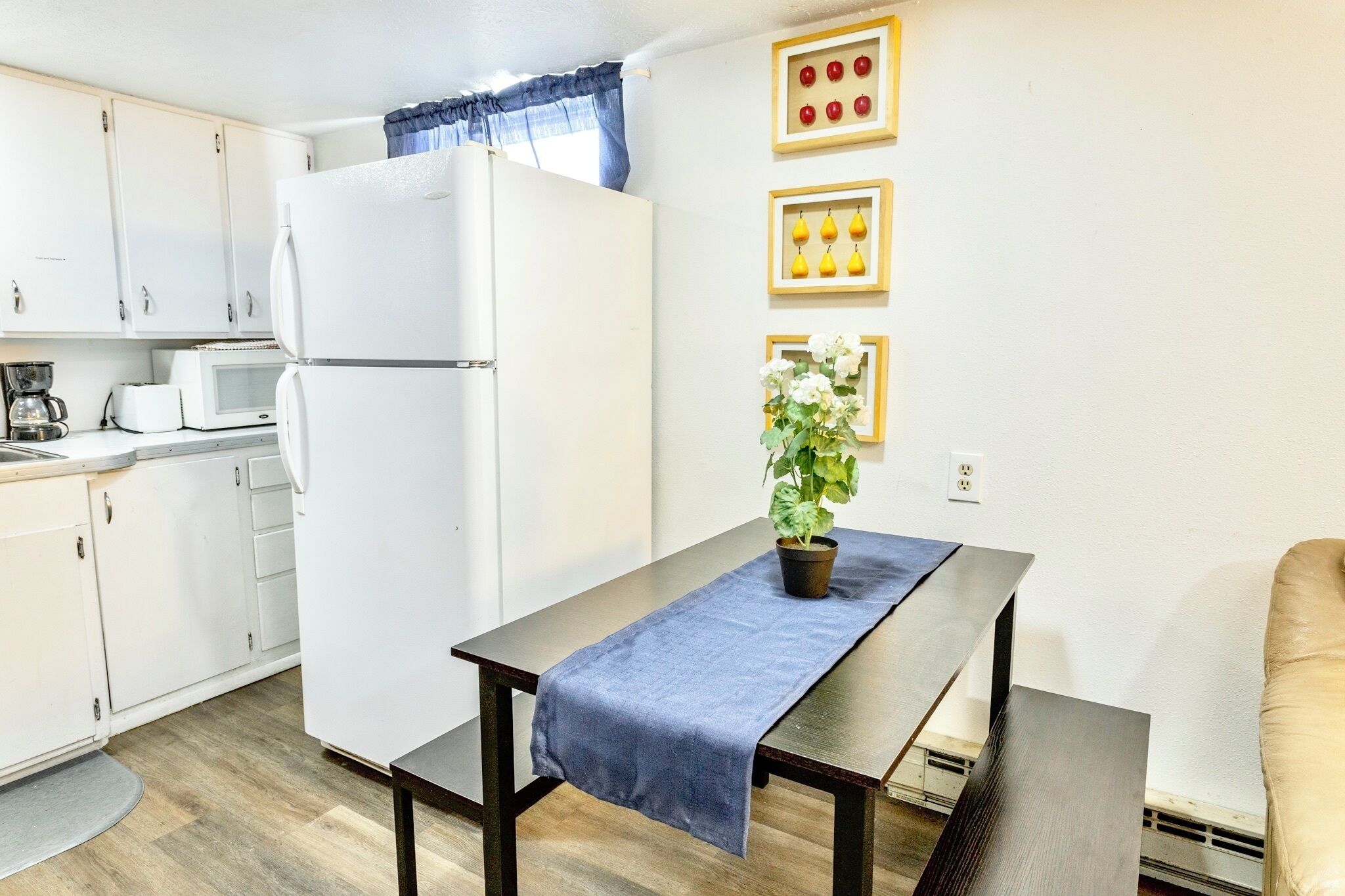 A compact kitchen with white cabinets, a white refrigerator, a microwave, and a coffee maker. There is a dining table with a blue tablecloth and a potted plant.