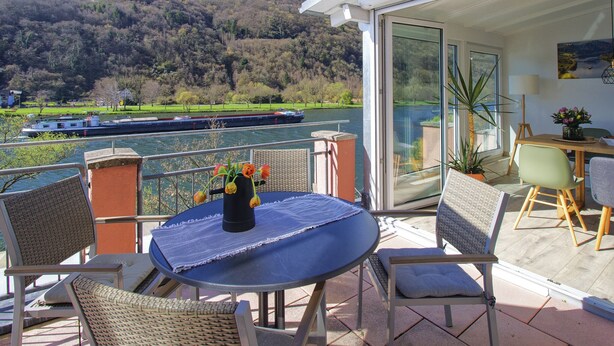 Un balcon avec des fauteuils en osier, une table ornée d’un vase de fleurs, une vue sur une rivière et un bateau aperçu au loin.