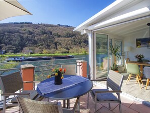 Ein Balkon mit Korbstühlen, einem Tisch mit Blumenvase und Blick auf einen Fluss, sowie ein Boot in der Ferne.
