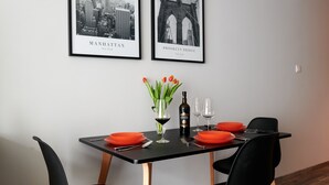 A dining area with a black table, four black chairs, a vase with tulips, wine glasses, and a bottle of wine. Two framed pictures of Manhattan and Brooklyn Bridge are on the wall. - Easy Rent Apartments - CRYSTAL (Lublin)