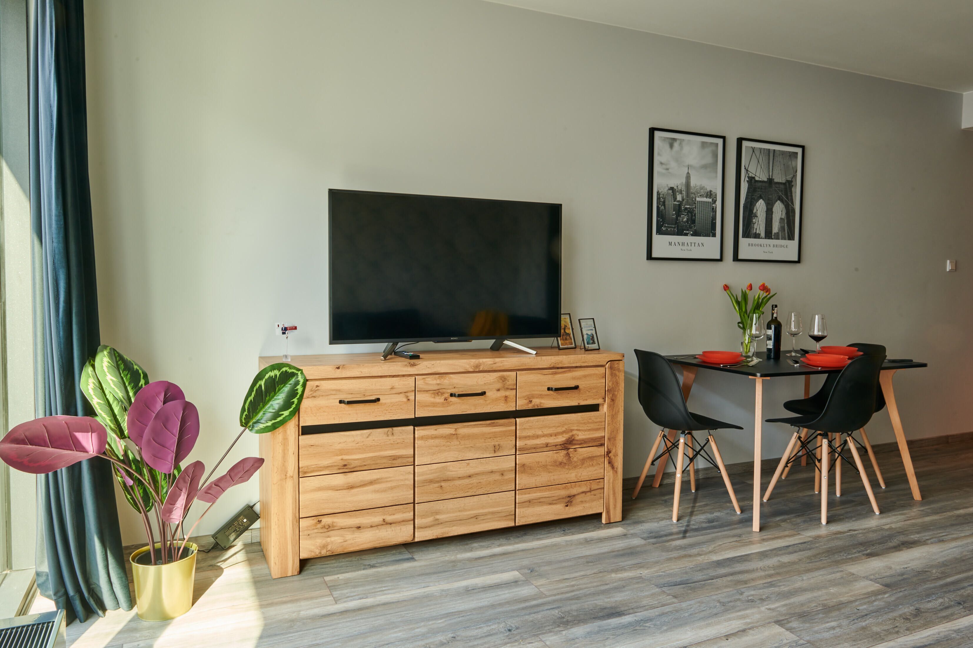 A modern living room with a wooden cabinet, a flat-screen TV, a dining table with chairs, and a potted plant.
