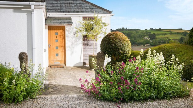Casa blanca con puerta de madera, rodeada por un camino de grava y diversas plantas.