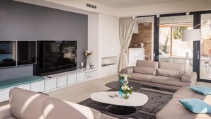 Una sala de estar moderna con un televisor de pantalla plana grande, sofás color beige, una mesa de centro y vistas al exterior a través de amplios ventanales.