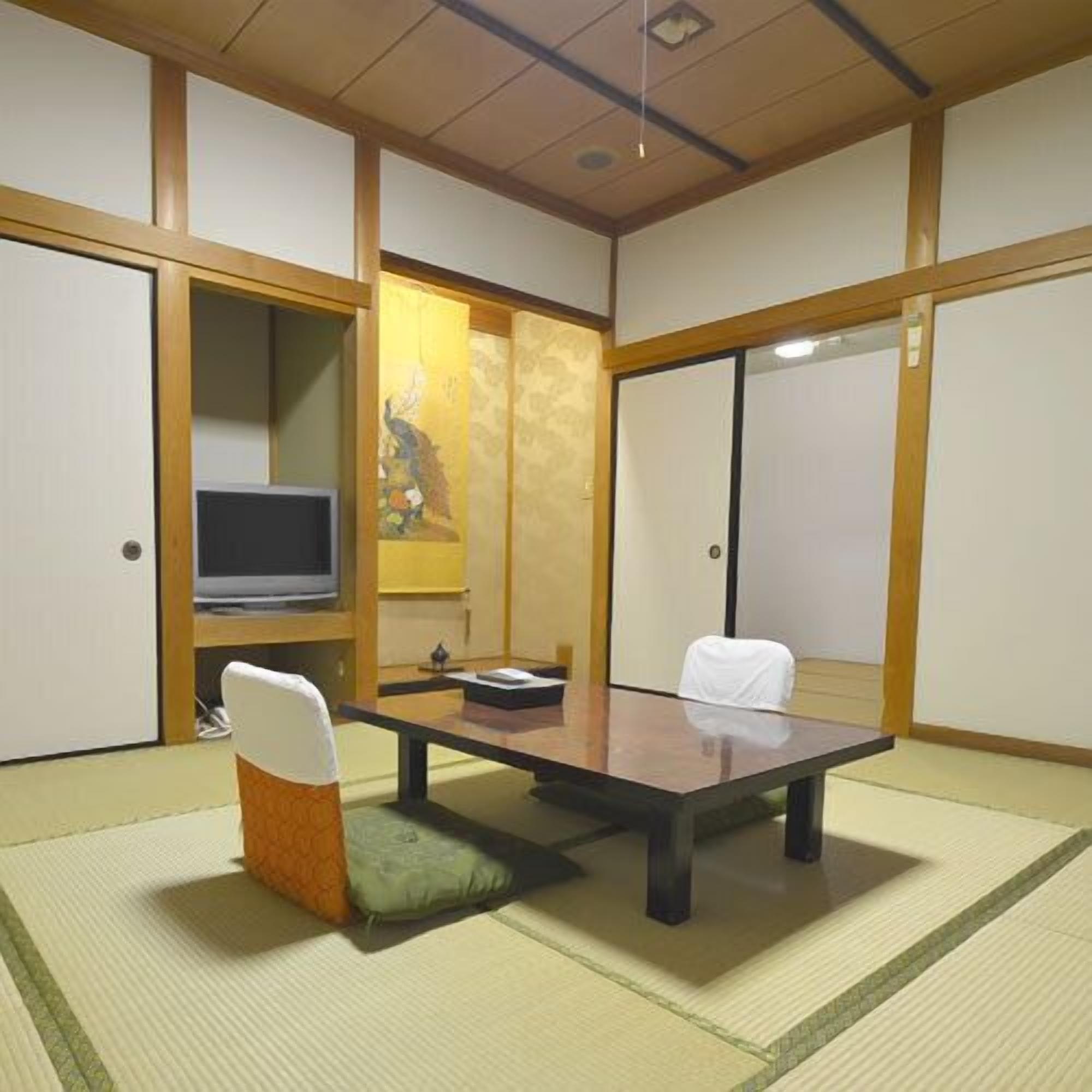 Una habitación japonesa tradicional con suelo de tatami, una mesa baja, dos sillas, un televisor y puertas correderas.