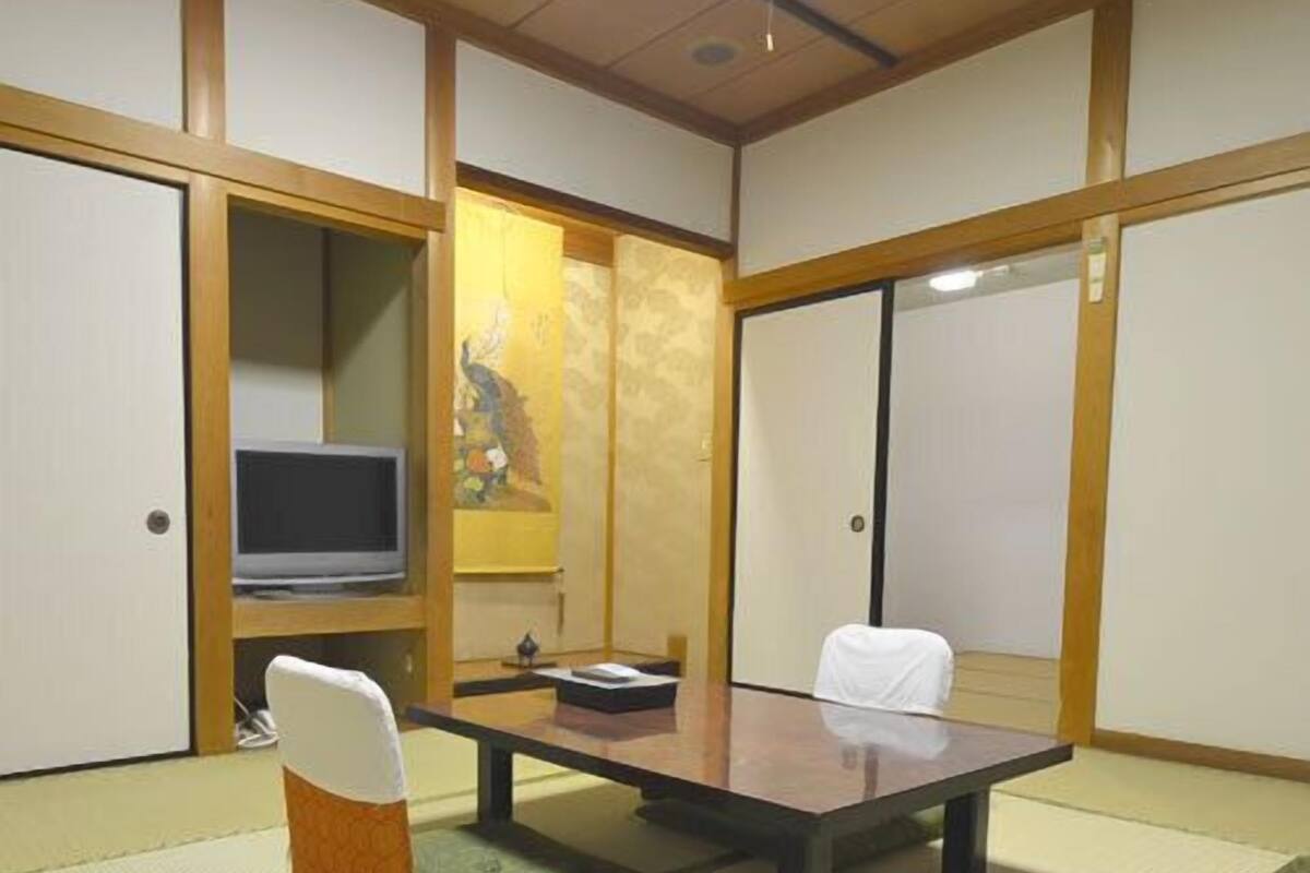 Una habitación japonesa tradicional con suelo de tatami, una mesa baja, dos sillas, un televisor y puertas correderas.