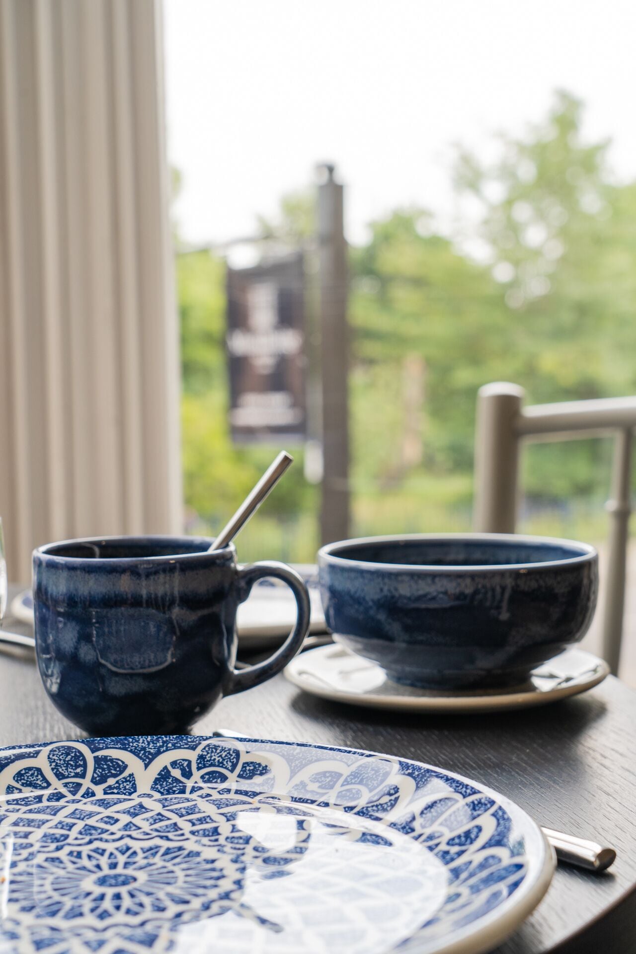 Una mesa con una taza de cerámica azul y un cuenco, un plato estampado y una silla.