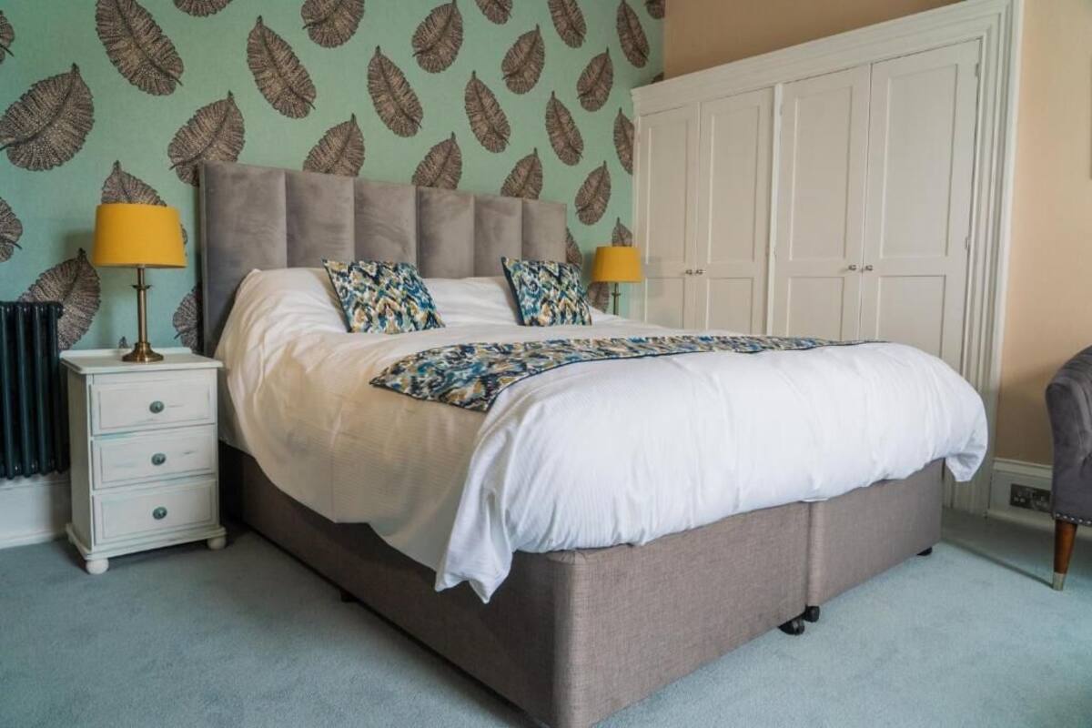 A bedroom with a large bed, two bedside tables, a wardrobe, and a patterned leaf wallpaper.