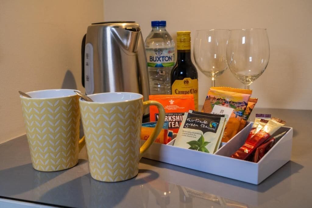 Una mesa con dos tazas amarillas, una botella de agua Buxton, una bolsita de té Campo Viejo, una cafetera y dos copas de vino vacías.