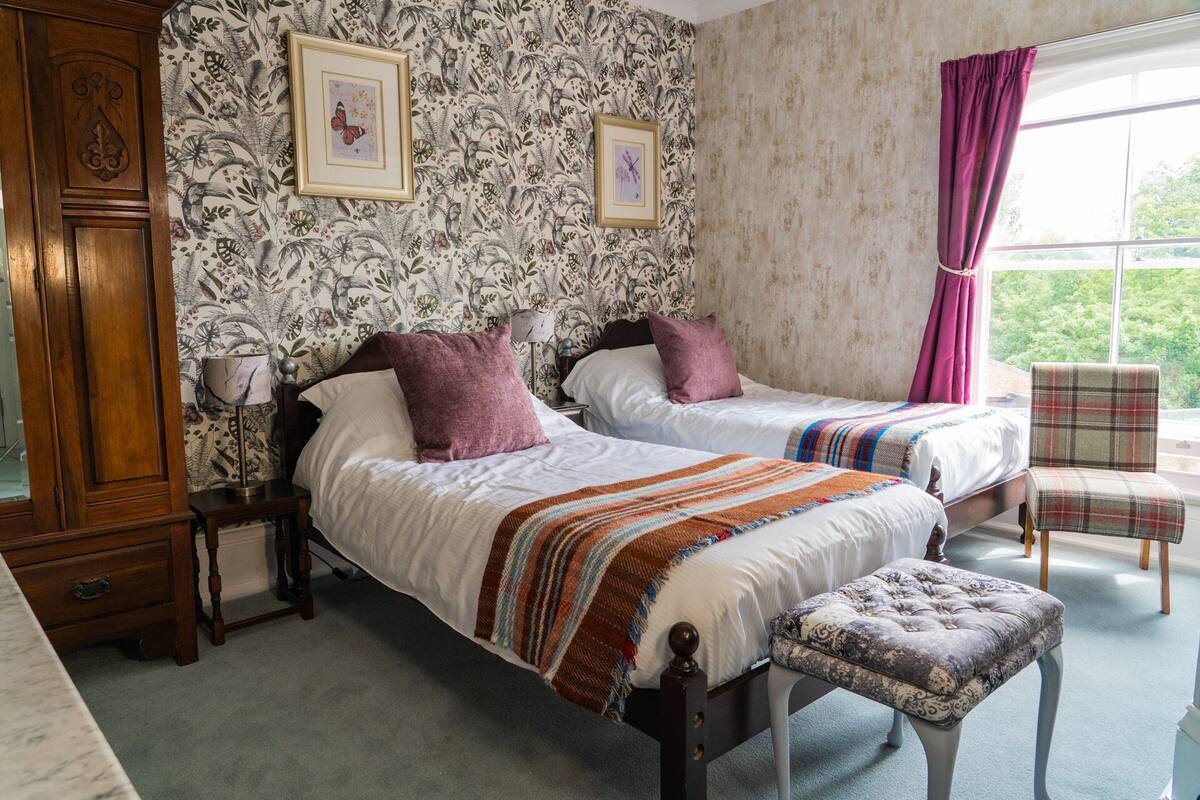 A bedroom with two beds, a wooden wardrobe, a chair, and a window with curtains.