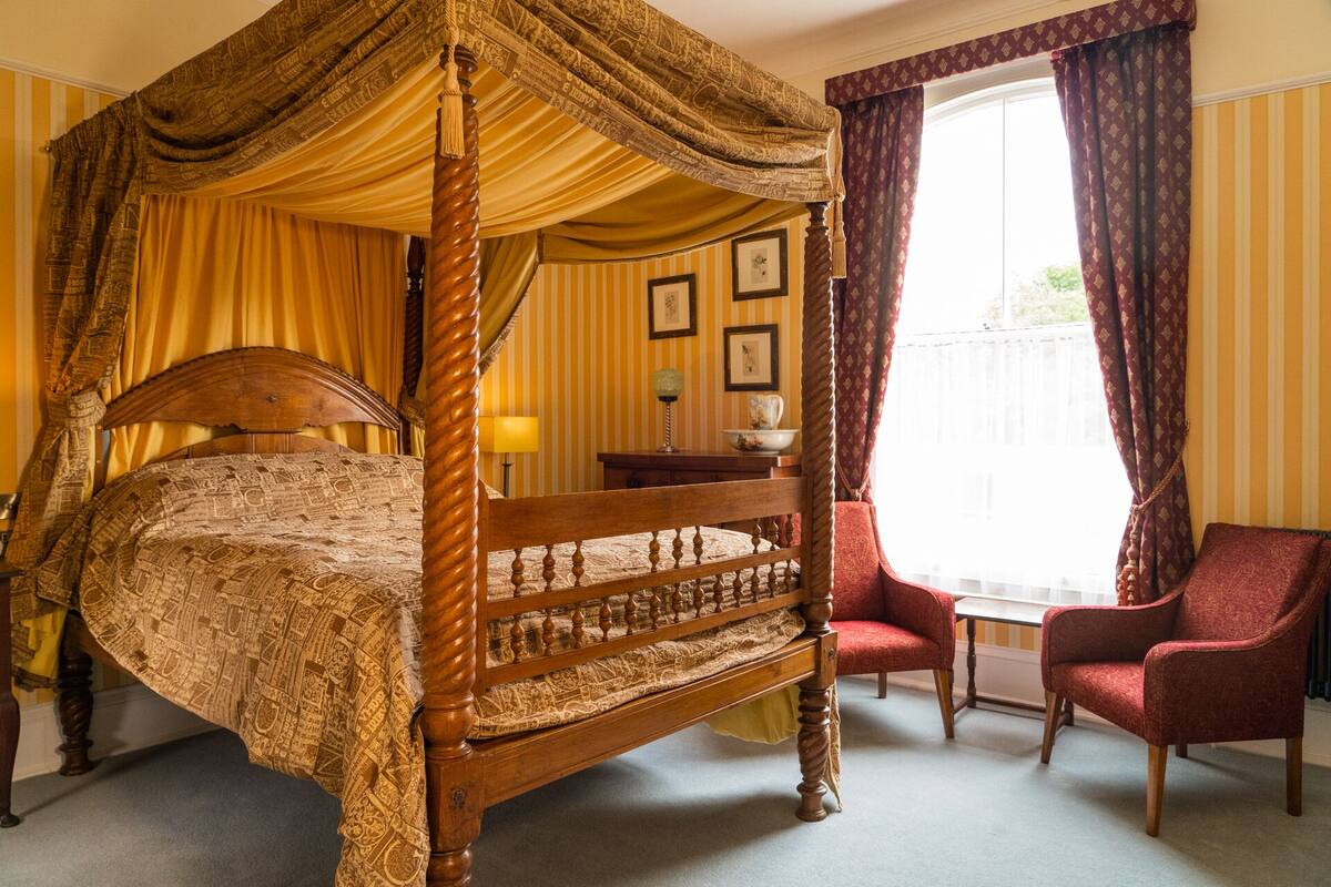 A four-poster canopy bed with a wooden frame and a patterned bedspread, a small table with a lamp, two red armchairs, and a window with curtains.
