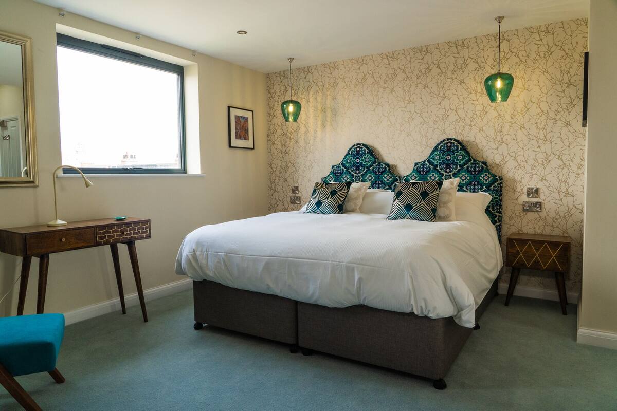 A bedroom with a large bed, a desk, and a chair. There is a window with a view of the outside.