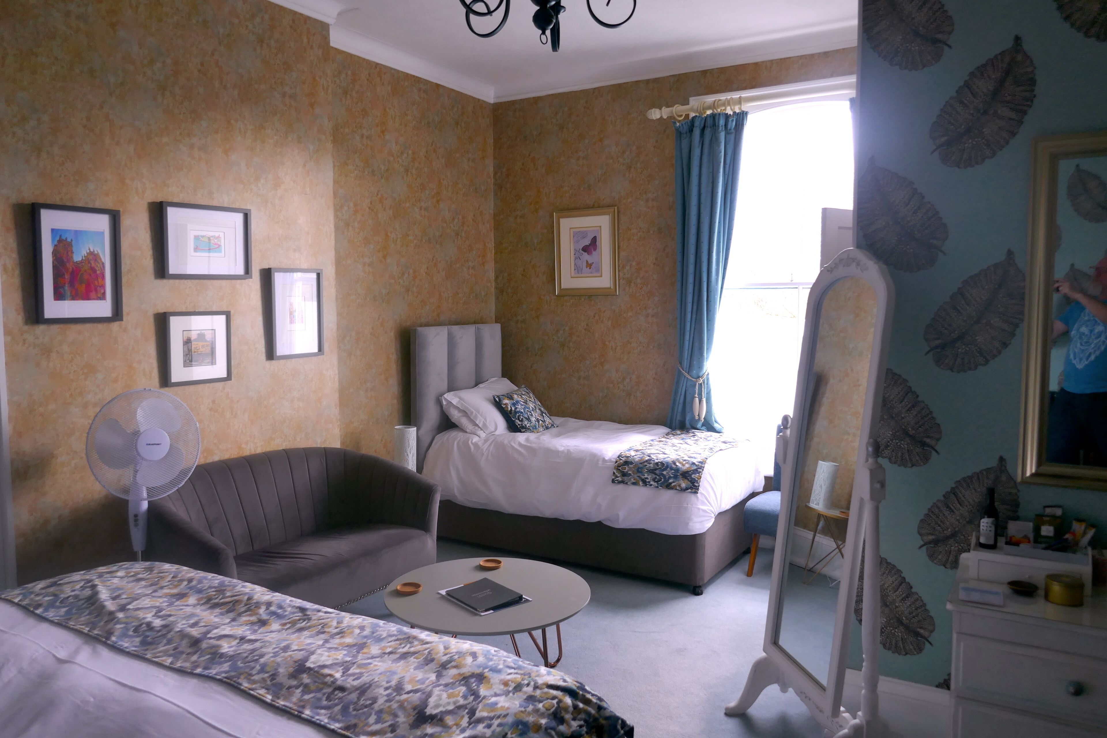 Une chambre d’hôtel avec un lit, une chaise, une petite table ronde, un ventilateur et un miroir.