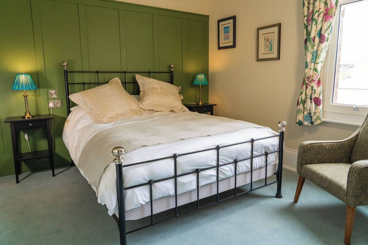 A neatly made bed with a metal frame, two bedside tables with lamps, a chair, and a window with floral curtains.