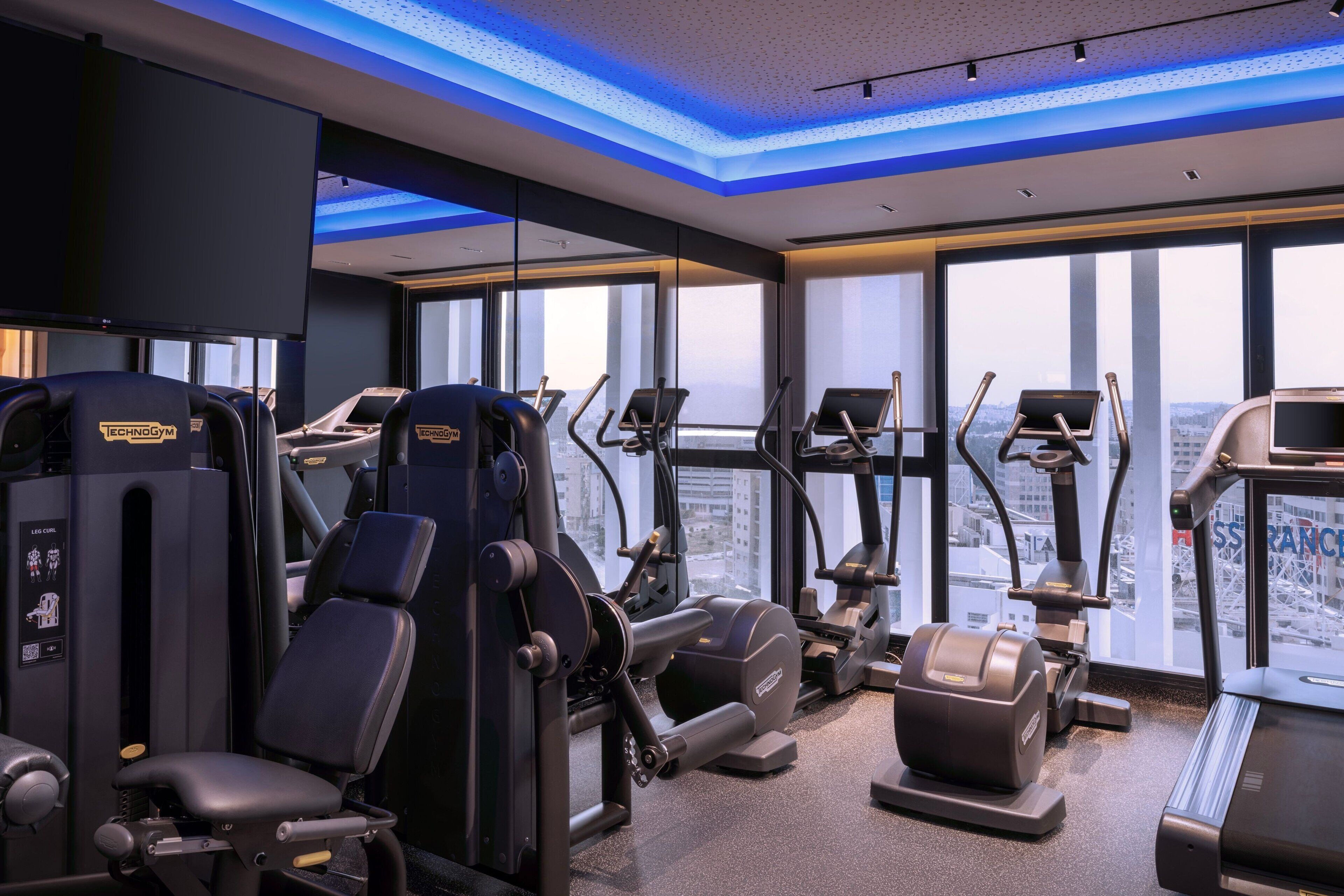 A modern gym with various exercise equipment, large windows offering city views, and a ceiling with blue lighting accents.