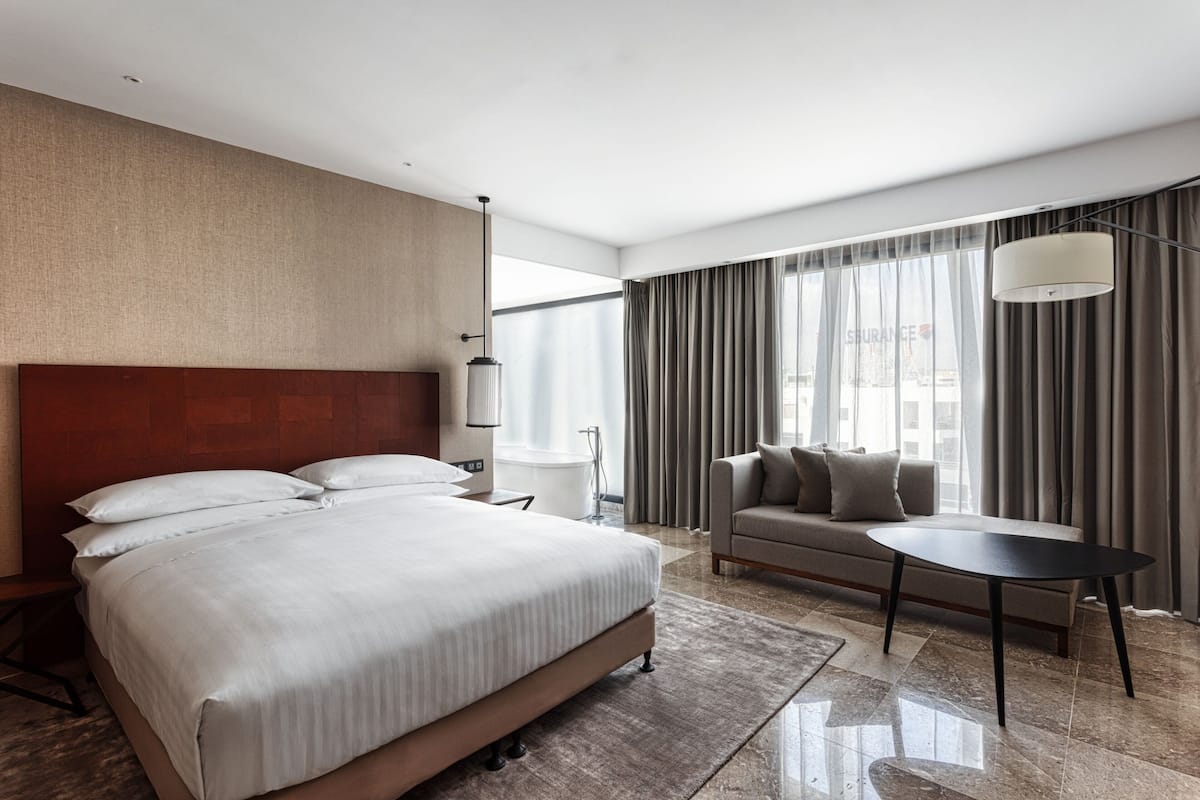 A modern hotel room with a large bed, a sofa, a small table, and a large window with curtains.