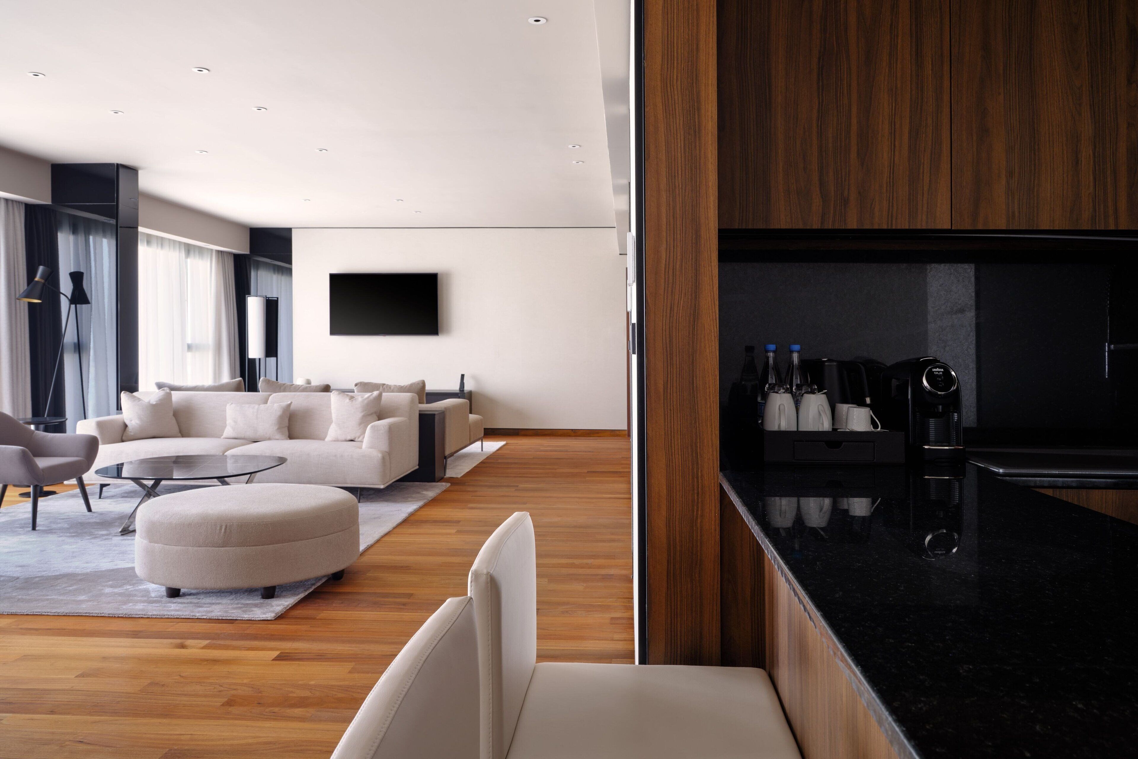 Un salon moderne avec un canapé, un pouf et une table basse. On y trouve également un coin cuisine doté d’un plan de travail noir et d’appareils électroménagers.