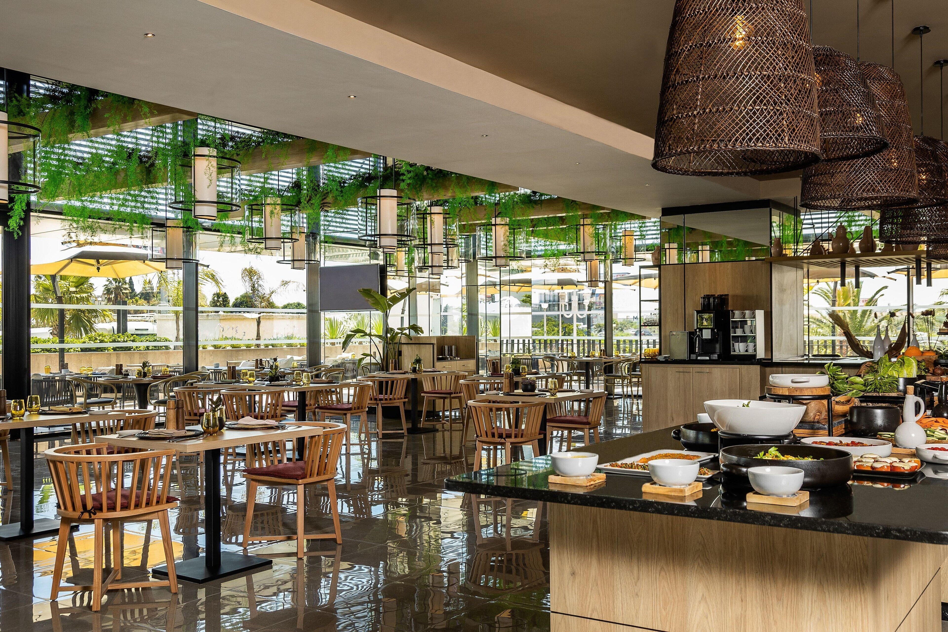 Un restaurante espacioso con sillas y mesas de madera, una isla central con comida y grandes ventanales con plantas.
