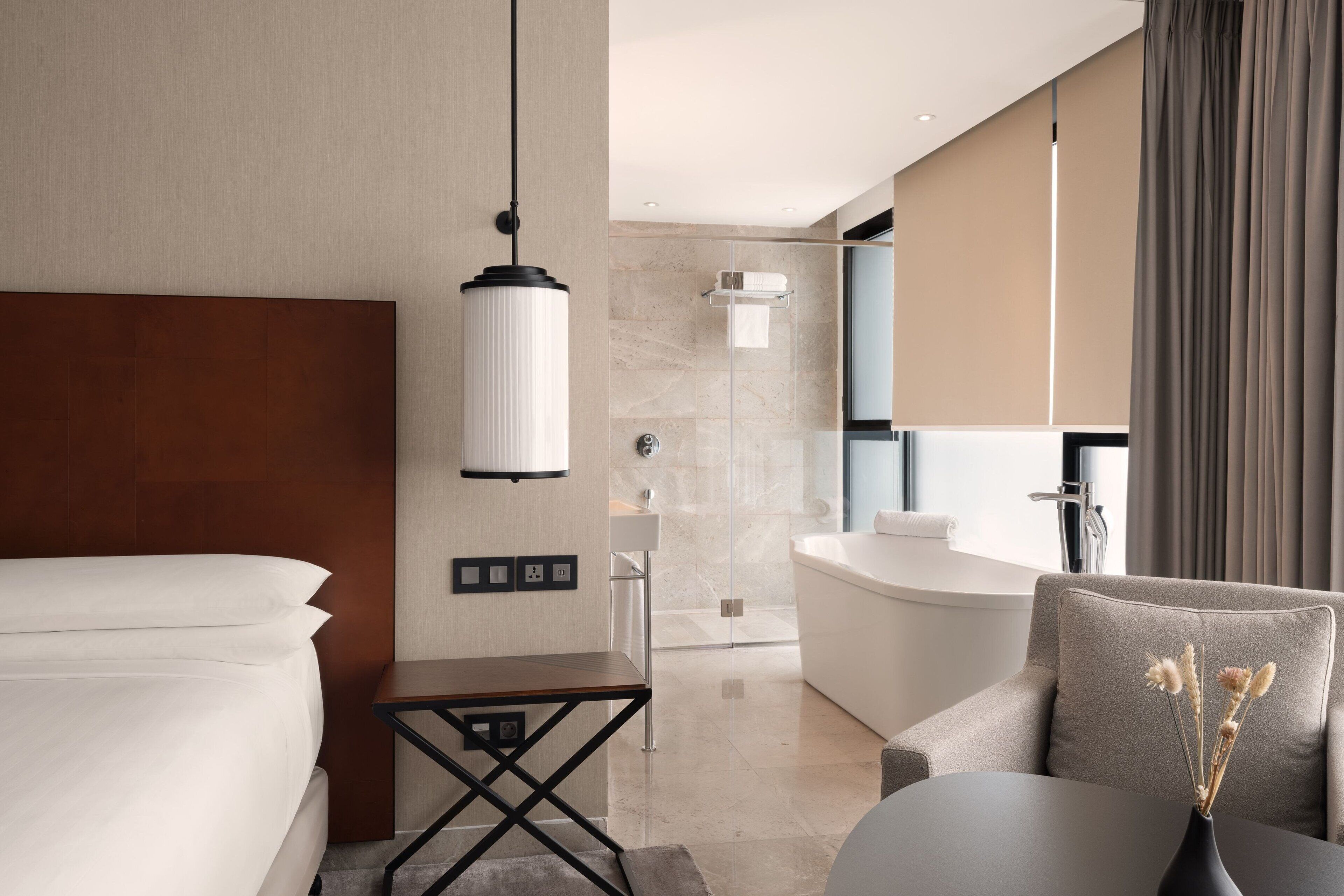 Ett modernt hotellrum med en säng, ett litet bord, en soffa, ett badrum med badkar och ett stort fönster med gardiner.