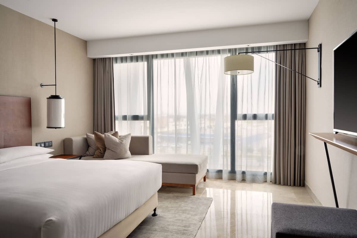 A modern hotel room with a large bed, a sofa, a TV, and a view of the city through large windows.