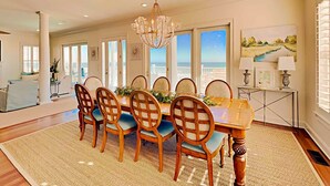 Une salle à manger avec une grande table, six chaises, un lustre et une vue sur l’océan grâce à de grandes fenêtres.