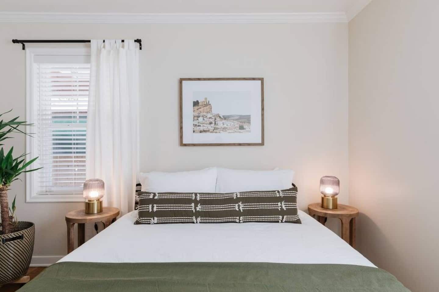 A neatly arranged bedroom with a bed, two bedside tables, a framed picture on the wall, and a window with curtains.