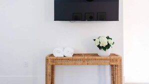 Ein Zimmer mit einem an der Wand befestigten Flachbildfernseher, einem Korbtisch mit gerollten Handtüchern und einer Blumenvase.