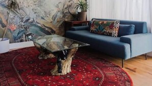 A living room with a blue sofa, a glass coffee table, a red patterned rug, a ceiling fan, and a mural of tropical foliage. - | The Jungle Bungalow | Downtown Chico | (Chico)