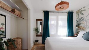 A bedroom with a bed, a hanging light fixture, a window with curtains, and a wall with a tropical mural. - | The Jungle Bungalow | Downtown Chico | (Chico)