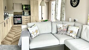 En moderne stue med en grå hjørnesofa, et køkken med træskabe, og et stort vindue med gardiner.